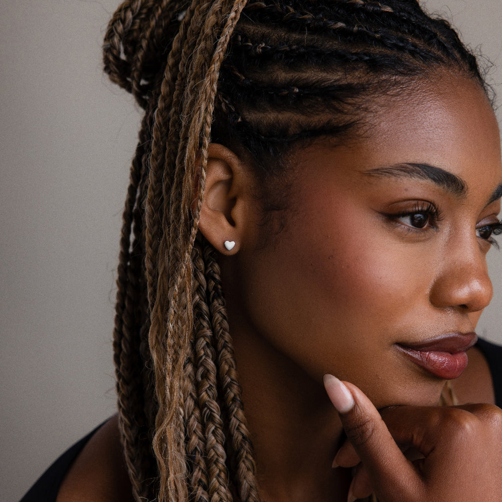 Woman with braided hair and a silver heart stud earring touching her face against a neutral background