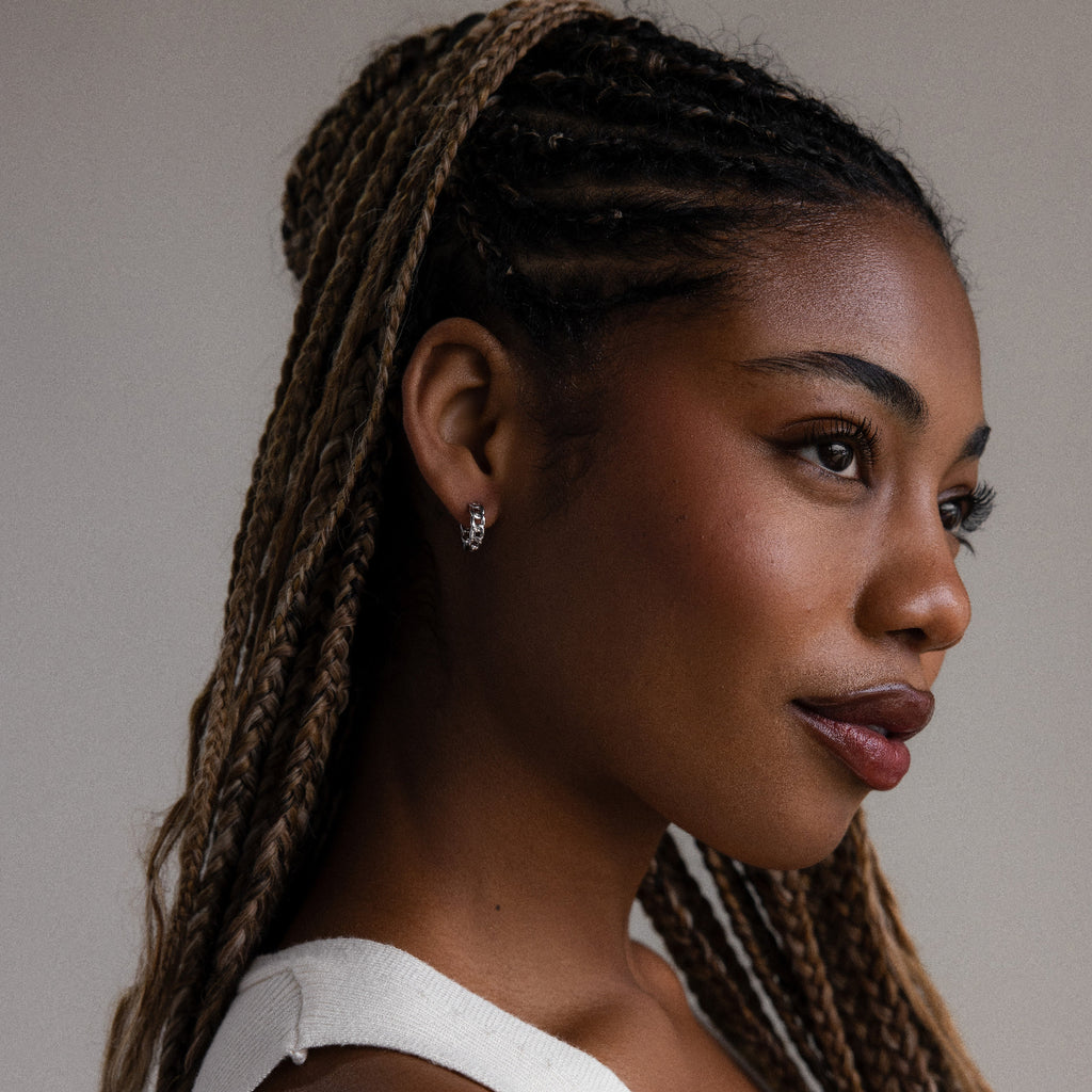 Woman with braided hair and silver chain hoop earrings wearing a white top against a neutral background
