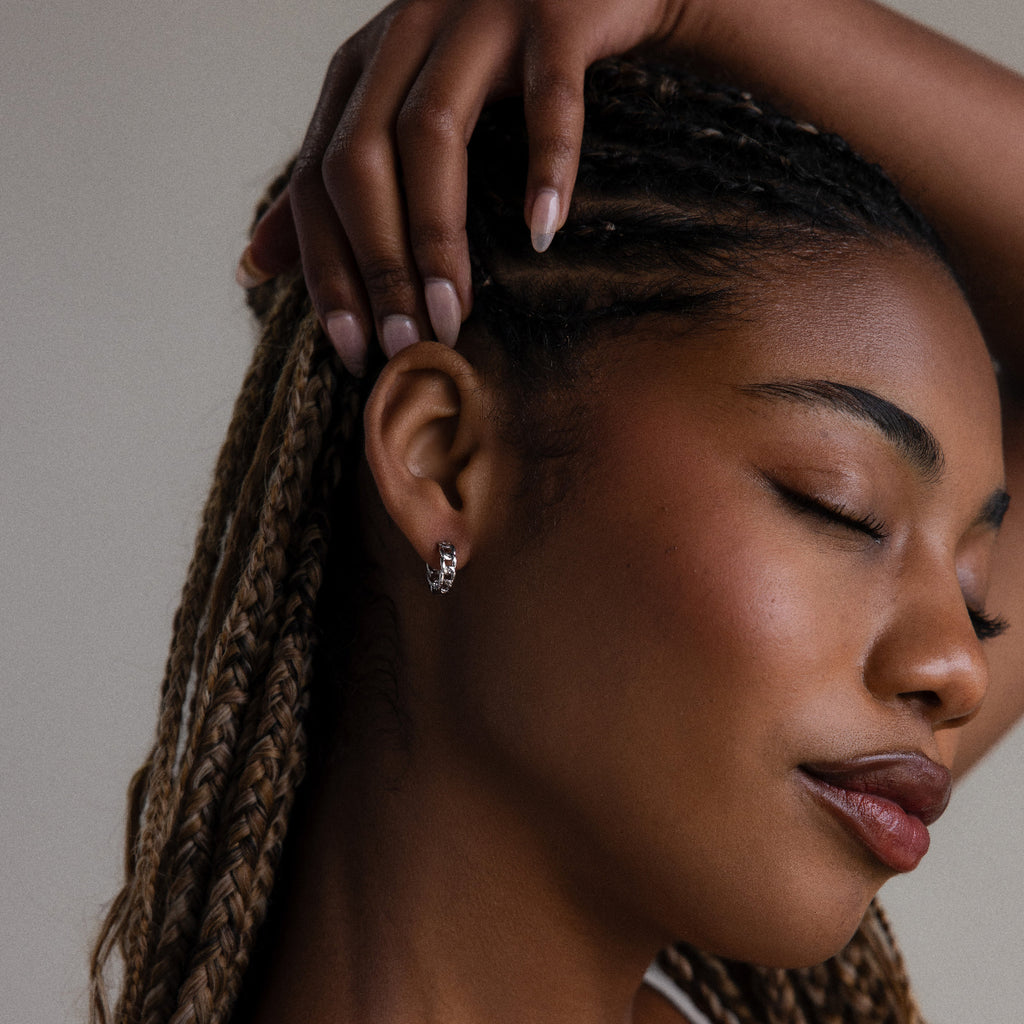 Close-up of a woman with braided hair and silver chain hoop earrings and her hand gently touching her head against a neutral background