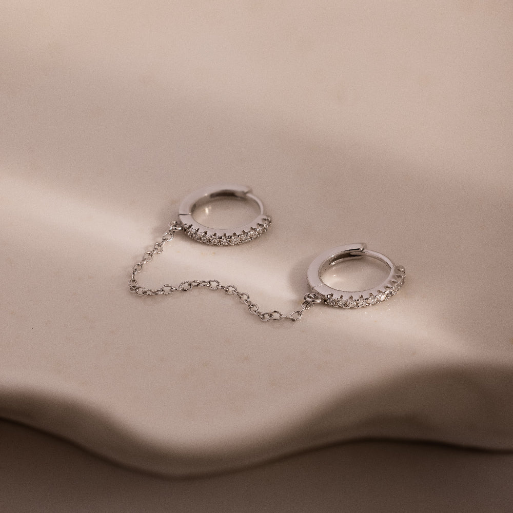 Pair of silver diamond hoop earrings with a chain on a beige surface