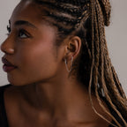 Close-up of a woman with braided hair wearing silver double diamond hoop earrings against a neutral background