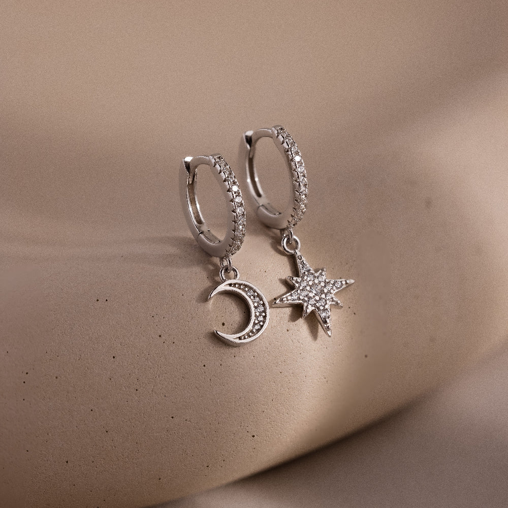 Silver hoop earrings with star and moon charms on a beige surface