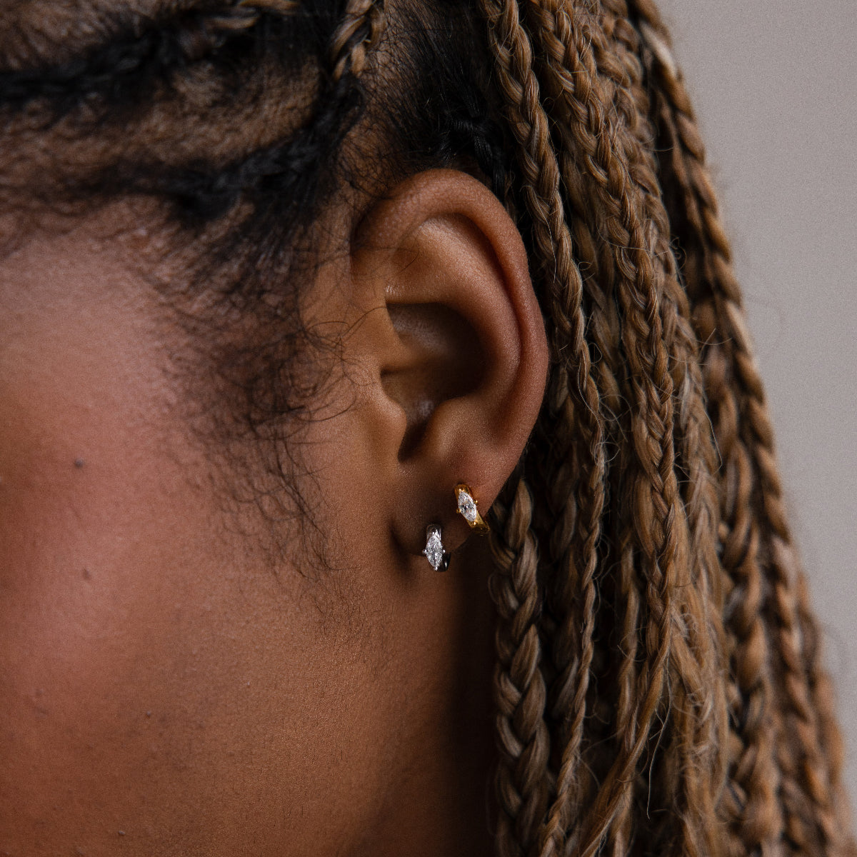 Close-up of a model with braided hair and gold and silver huggie hoop earrings against a neutral background