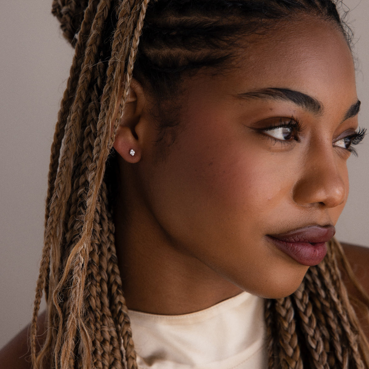 Close-up of a woman with braided hair wearing a silver diamond stud earring and a white top against a neutral background