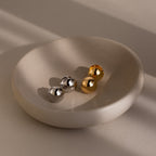 Four metallic sphere earrings in gold and silver on a beige ceramic dish with a neutral background