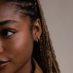 Close-up of a person with braided hair wearing silver and gold baguette hoop earrings against a neutral background