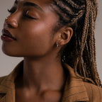 Woman with braided hair wearing a silver hoop earring and a brown jacket against a neutral background