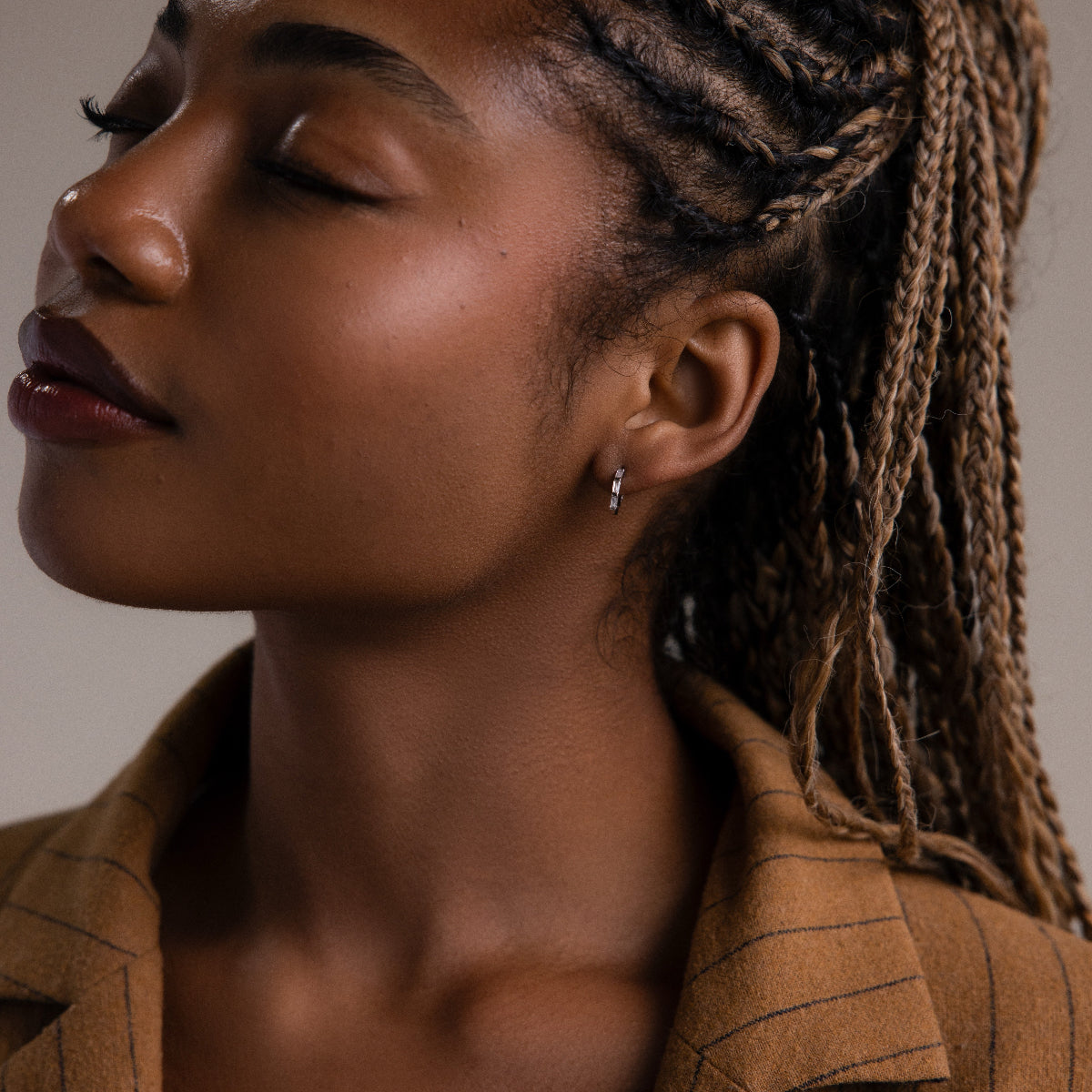 Woman with braided hair wearing a silver hoop earring and a brown jacket against a neutral background