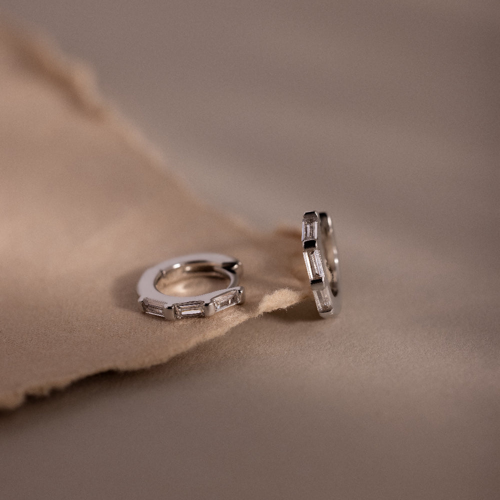Pair of silver hoop earrings with clear baguette cut gemstones on a soft brown fabric background