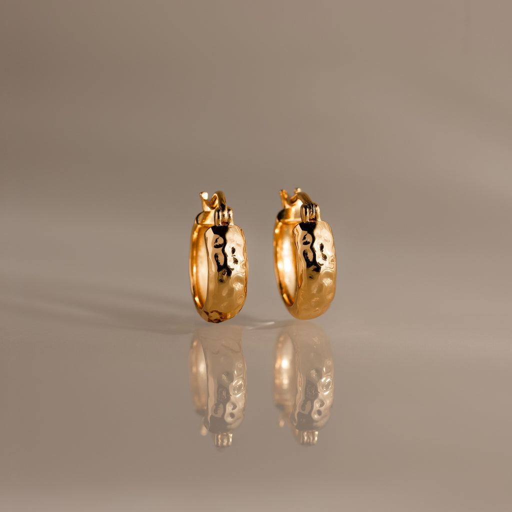 Bold Hammered Huggies in 18K Gold with a textured finish and abstract black designs are displayed on a reflective, neutral-colored surface.