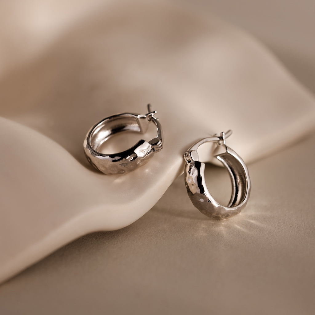 A pair of Bold Hammered Huggies in Silver with a textured finish rests on a smooth, light surface—ideal for fans of minimal design jewelry.