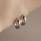 Bold Hammered Huggies in Silver feature a textured finish with small black accents and a minimal design, elegantly displayed on beige textured paper.
