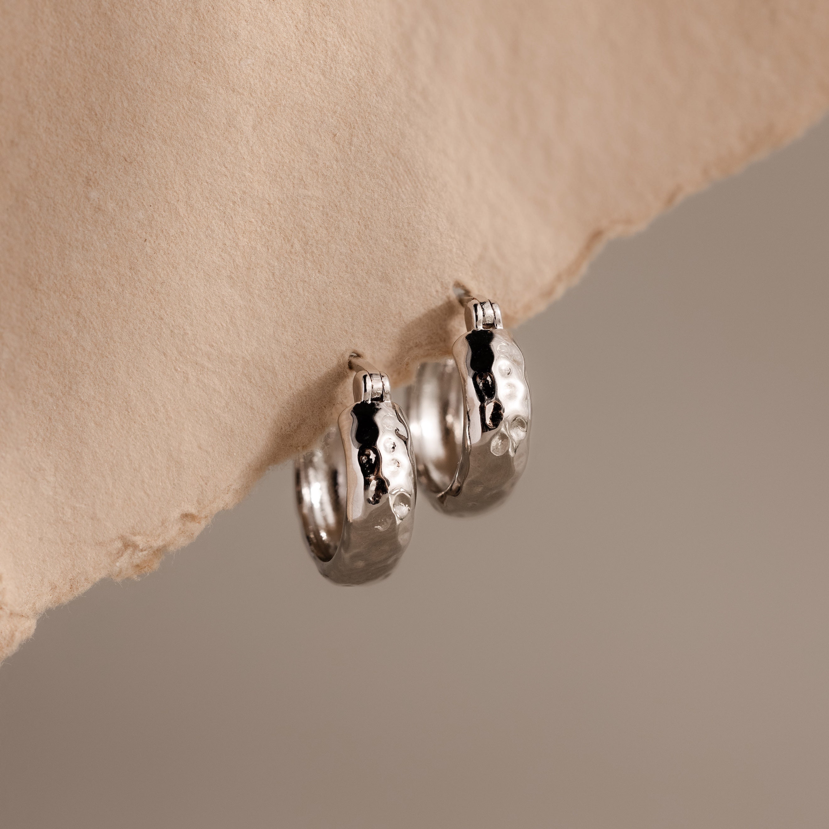 Bold Hammered Huggies in Silver feature a textured finish with small black accents and a minimal design, elegantly displayed on beige textured paper.