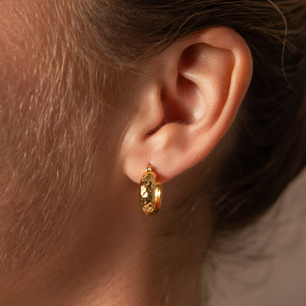 A close-up of an ear wearing Bold Hammered Huggies, highlighting this minimalist earring’s bold, textured gold finish.