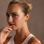 A blonde woman in a bun, wearing a white tank top and Bold Hammered Huggies in Silver, gazes thoughtfully with her hand near her chin.