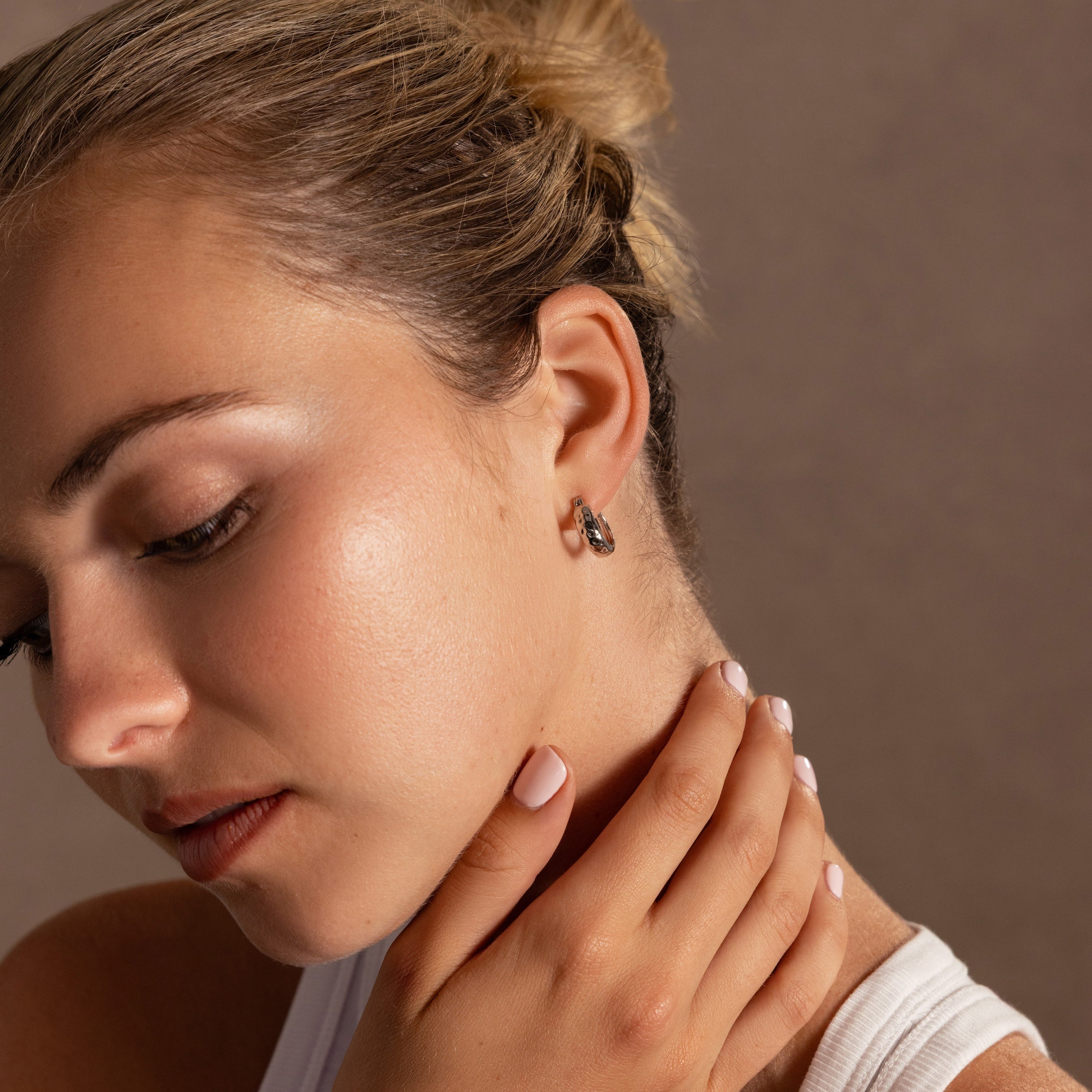 A woman with blonde hair in a bun touches her neck, wearing a white top and Bold Hammered Huggies in Silver that add a touch of minimal design to her look.
