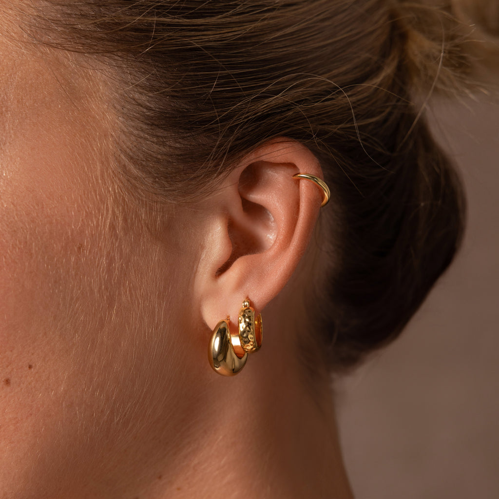 Close-up of a woman’s ear with minimal design jewelry—silver Bold Hammered Huggies, an ear cuff, and textured earrings, hair tied back.