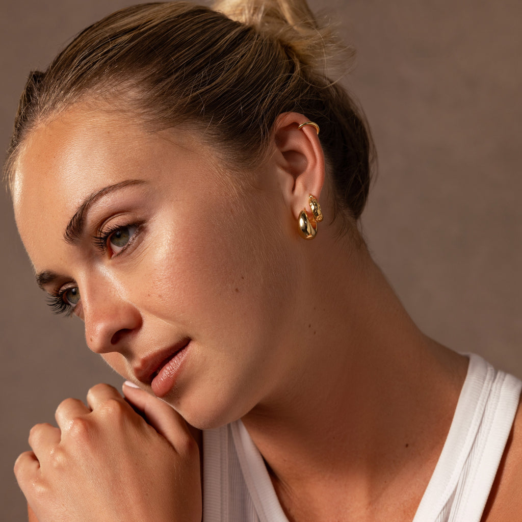 Blonde-haired woman with a bun, wearing a white top and Bold Hammered Huggies earrings with a textured finish, looks thoughtful.