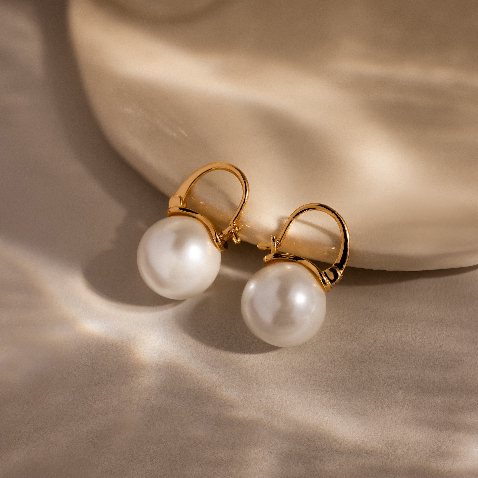 The Pearl Dome Drop Earrings feature lustrous pearls and a classic design, showcased on a smooth beige surface with soft lighting to highlight their timeless elegance.