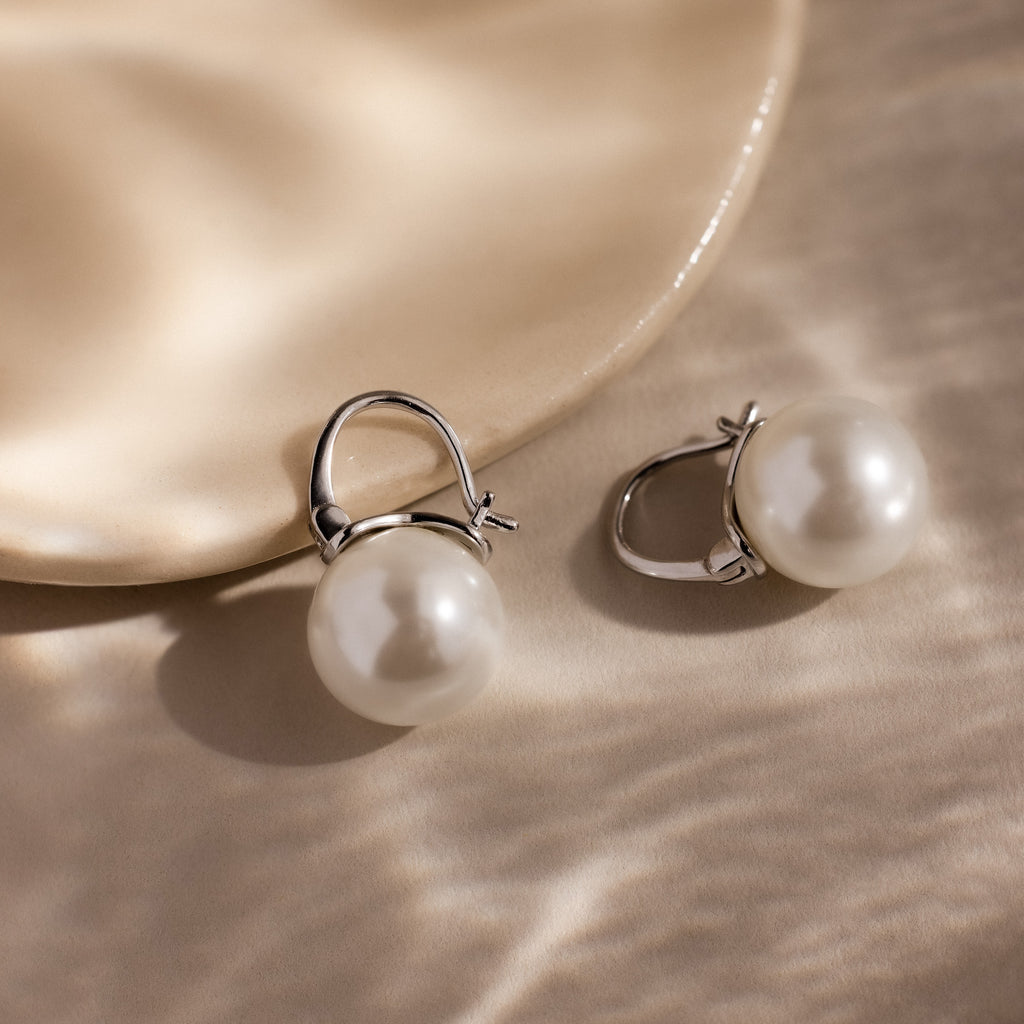 The Pearl Dome Drop Earrings, featuring lustrous pearls and silver clasps, rest elegantly on a smooth beige surface.