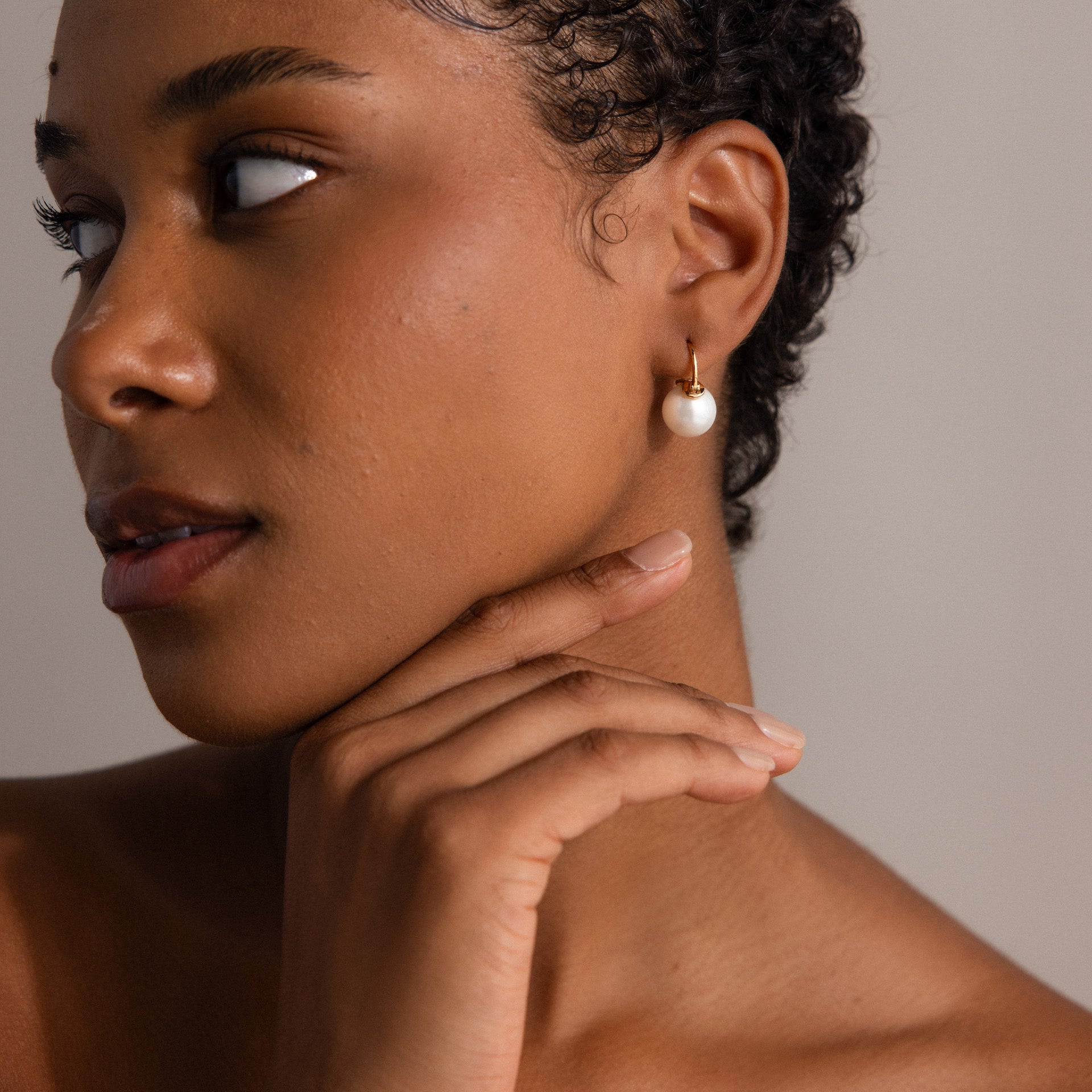 A woman with short curly hair wears Pearl Dome Drop Earrings and touches her chin, gazing to the side.