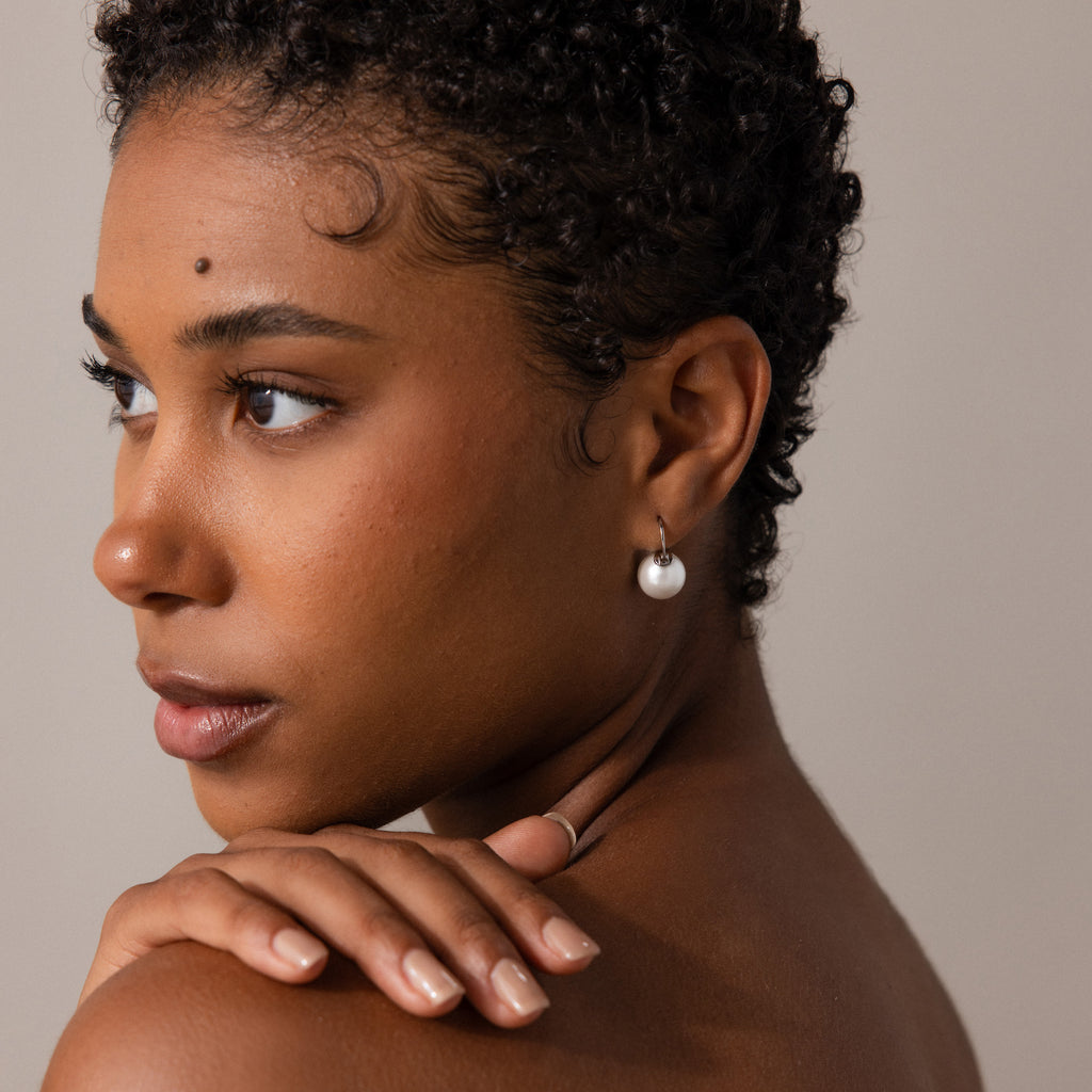 A woman with short curly hair wears Pearl Dome Drop Earrings, looking to the side with her hand on her shoulder against a neutral background.