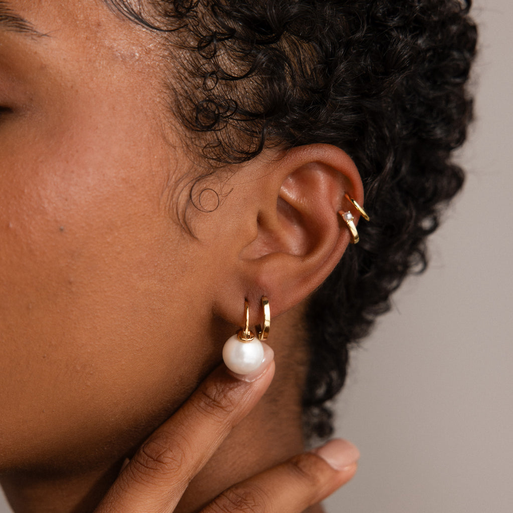 A close-up of an ear wearing elegant earrings, including a gold hoop and the Pearl Dome Drop Earrings with lustrous pearls, as a hand gently touches the earlobe.
