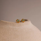 Close-up of gold flower-shaped stud earrings with a round green jade gemstone center, displayed on a neutral stone surface.