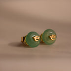 Close-up of gold Jade Bird Studs with dainty golden dove birds on round green jade discs, displayed on a neutral beige surface.