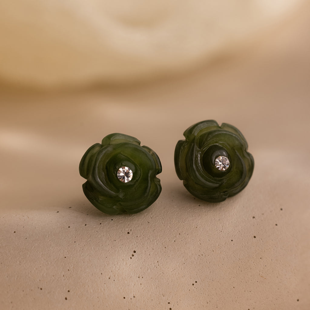 Close-up of the jade rose studs on a textured neutral surface, showing fine carved petals and crystal centers in detail.
