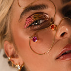 Close-up of a woman’s face with bold eye makeup, gold jewelry, and colorful pendants on her skin, featuring a striking Hexagon Birthstone Charm in a geometric frame.