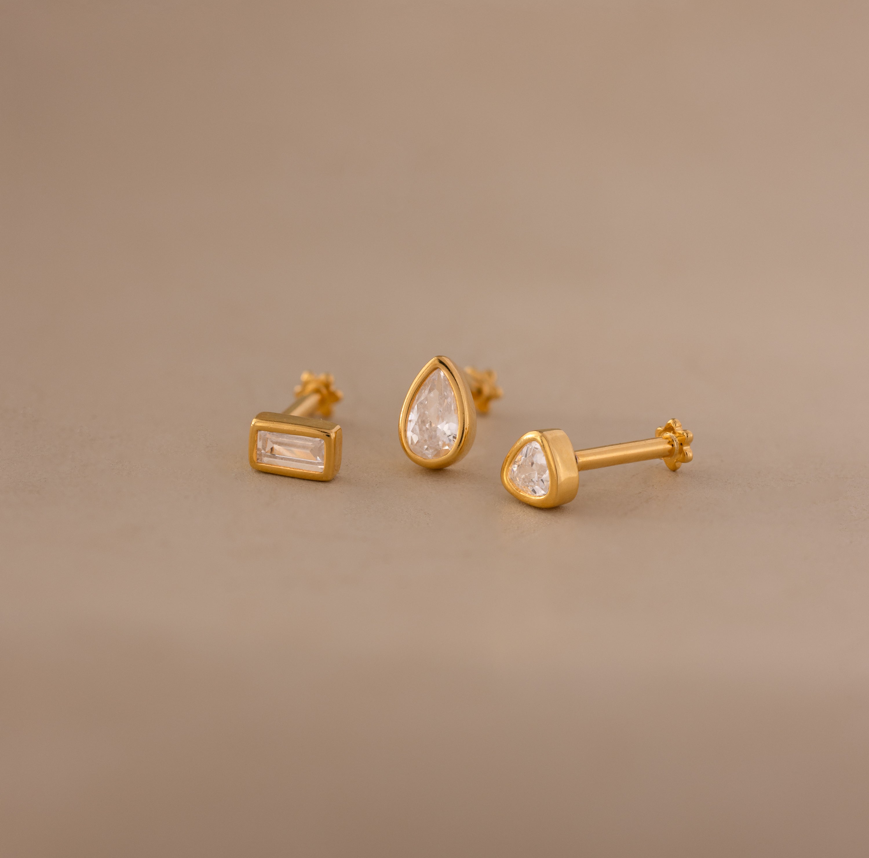 Three Diamond Bezel Flatback Studs in 18K Gold, each featuring a unique clear gemstone shape—rectangle, teardrop, and triangle—displayed on a beige background for a modern look.