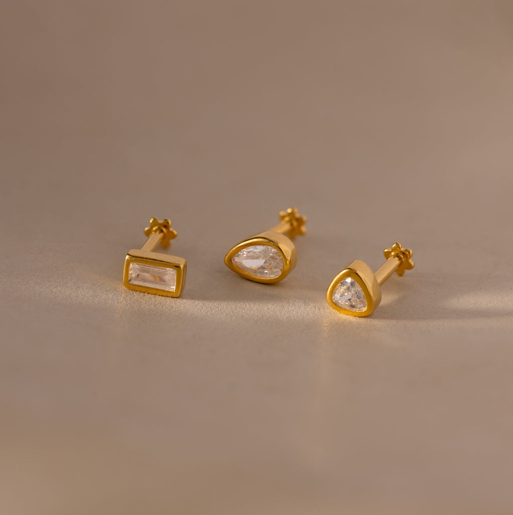Set of three Diamond Bezel Flatback Studs in 18K Gold, each with a unique geometric shape and clear gemstones, displayed on a beige surface.
