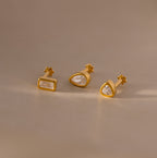Set of three Diamond Bezel Flatback Studs in 18K Gold, each with a unique geometric shape and clear gemstones, displayed on a beige surface.