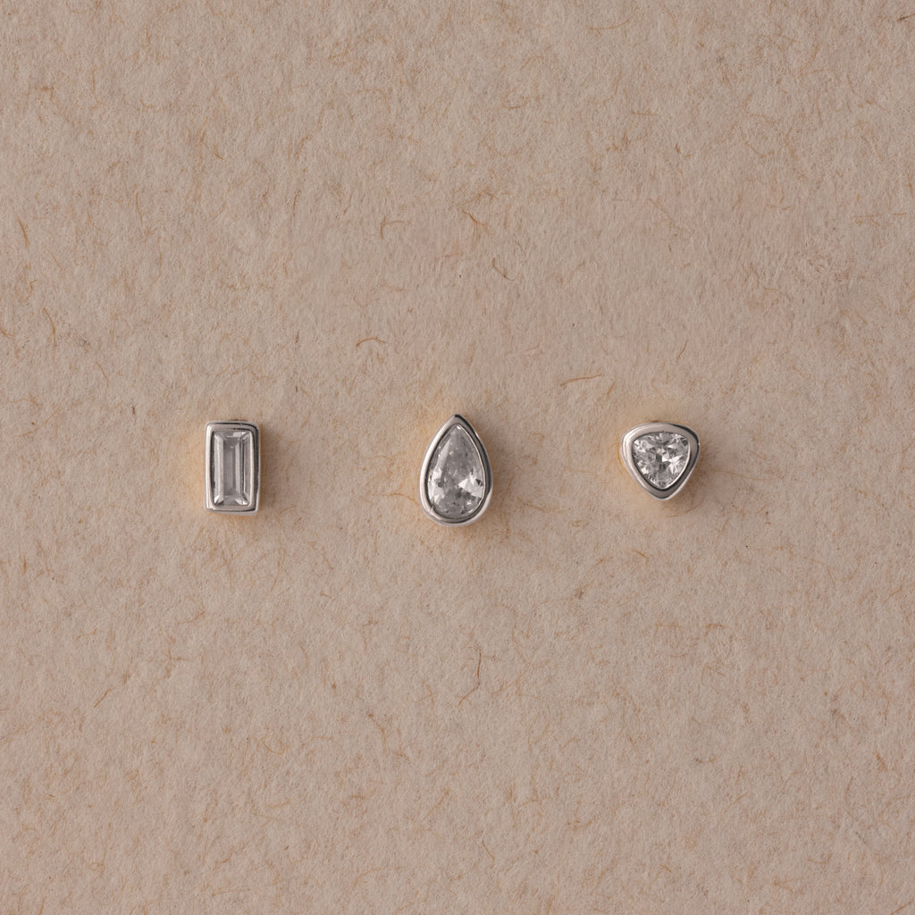 The Diamond Bezel Flatback Studs Set in Silver features rectangular, teardrop, and triangular gemstones on a beige textured background for a classic, elegant look.
