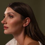 Woman with long brown hair wears Diamond Bezel Flatback Studs Set in 18K Gold, gazing left against a plain olive background.