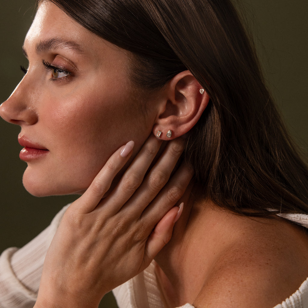 A woman with long nails touches her neck, highlighting Diamond Bezel Flatback Studs Set in Silver while wearing a cream-colored top against a dark background.