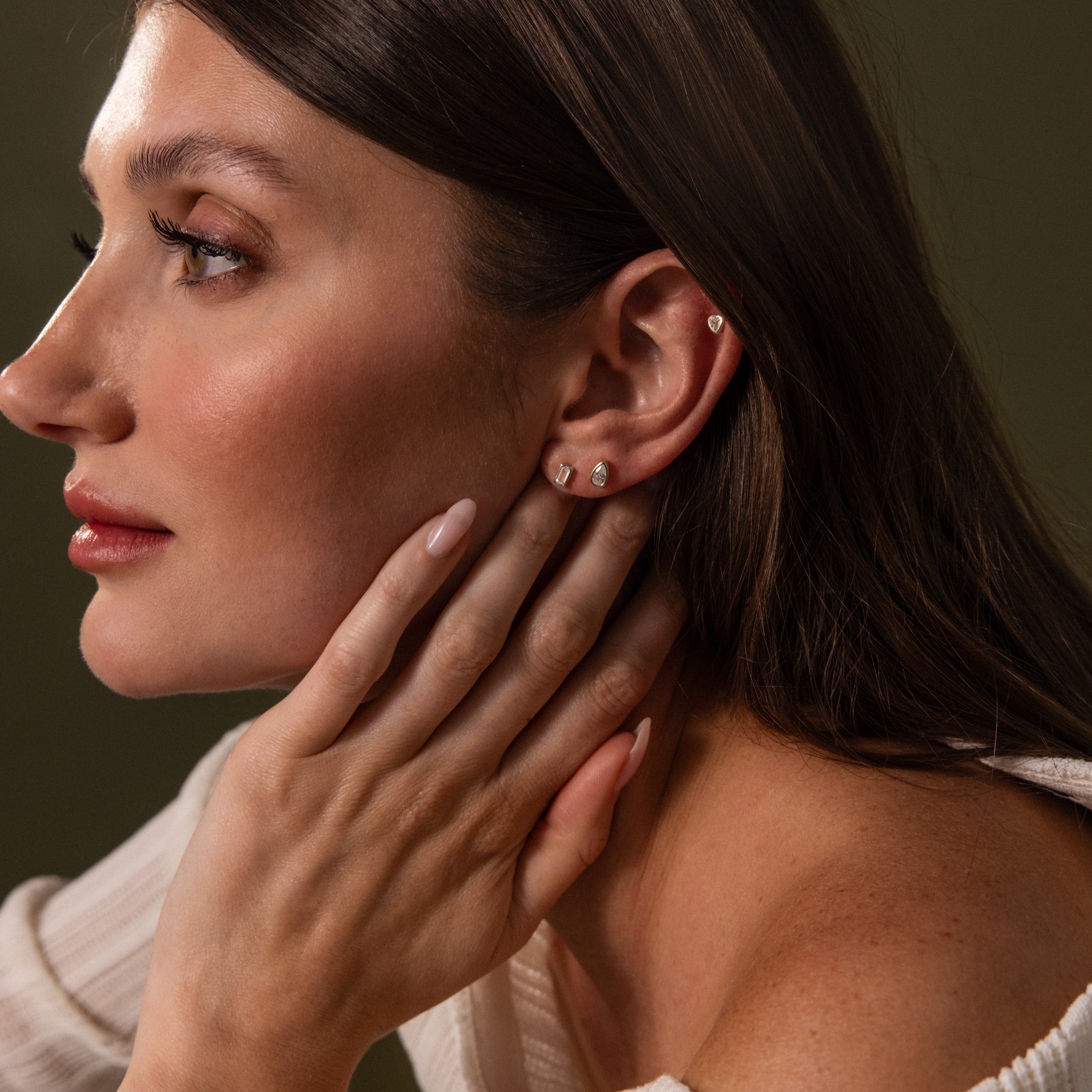 A woman with long nails touches her neck, highlighting Diamond Bezel Flatback Studs Set in Silver while wearing a cream-colored top against a dark background.