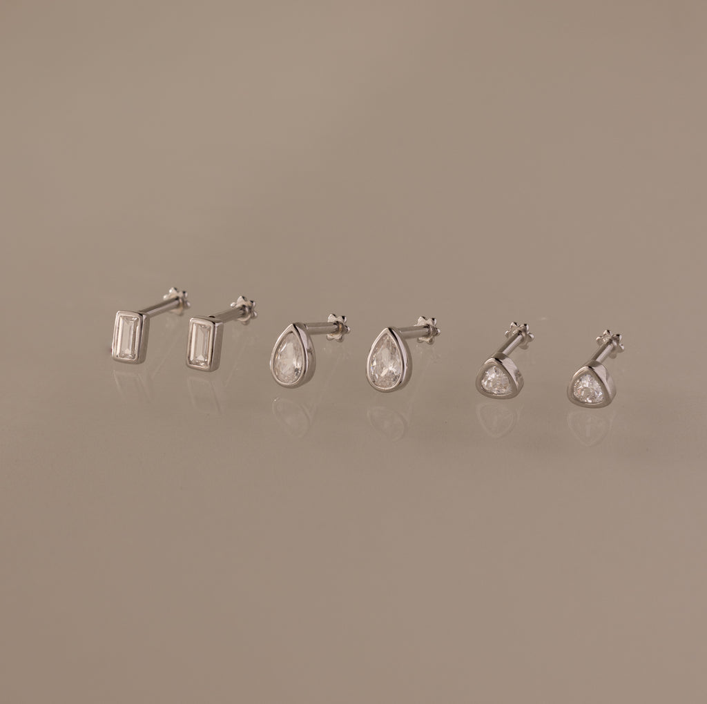 The Diamond Bezel Flatback Studs Set in Silver features three pairs of classic cut earrings—rectangular, teardrop, and round—elegantly displayed on a beige background.