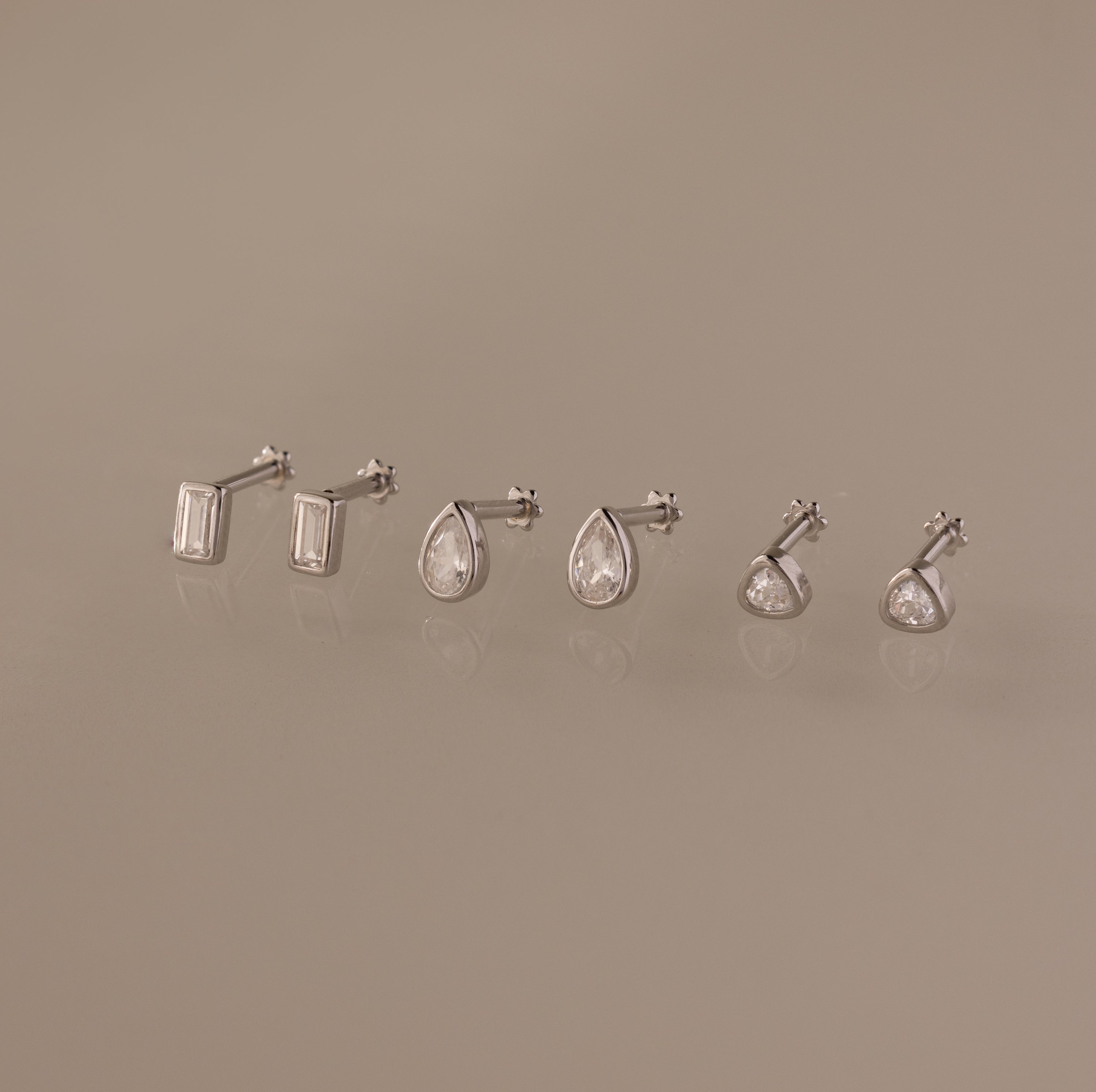 The Diamond Bezel Flatback Studs Set in Silver features three pairs of classic cut earrings—rectangular, teardrop, and round—elegantly displayed on a beige background.