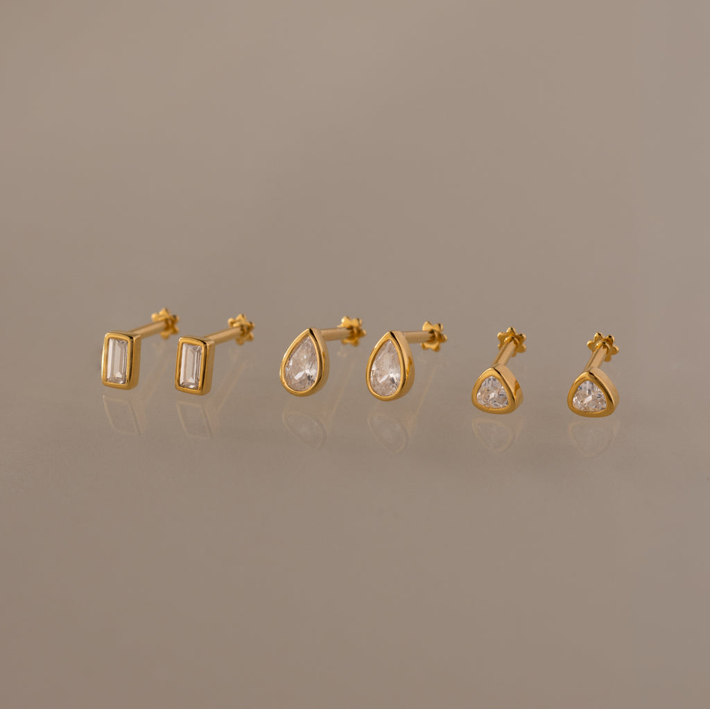 The Diamond Bezel Flatback Studs Set in 18K Gold features three pairs of gold stud earrings—rectangular, teardrop, and round shapes with clear stones—on a neutral background.