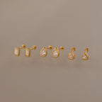 The Diamond Bezel Flatback Studs Set in 18K Gold features three pairs of gold stud earrings—rectangular, teardrop, and round shapes with clear stones—on a neutral background.