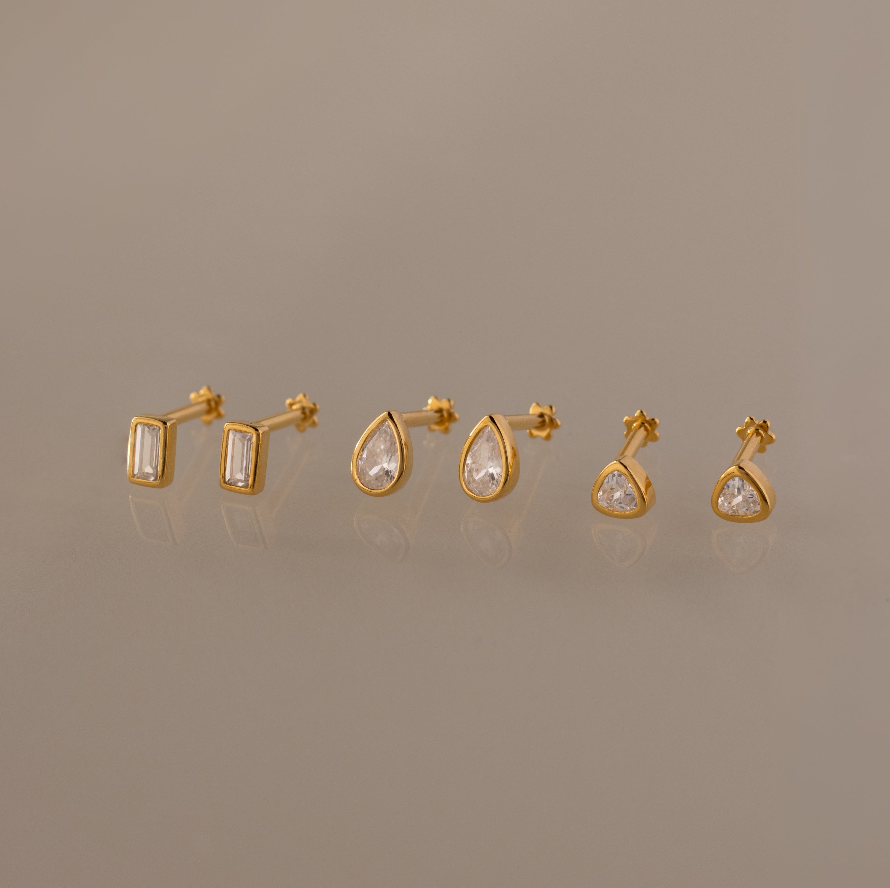 The Diamond Bezel Flatback Studs Set in 18K Gold features three pairs of gold stud earrings—rectangular, teardrop, and round shapes with clear stones—on a neutral background.