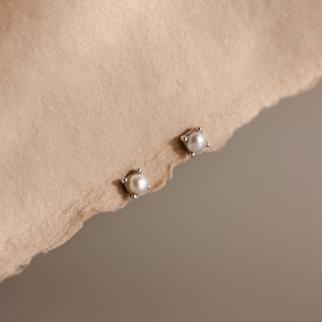 Two Baby Venus Flatback Studs in Silver, featuring delicate pearl earrings, are displayed on a soft beige fabric surface.