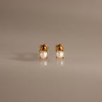 A pair of Baby Venus Flatback Studs in 18K Gold, featuring delicate pearls and floral backing, displayed on a smooth, reflective surface.