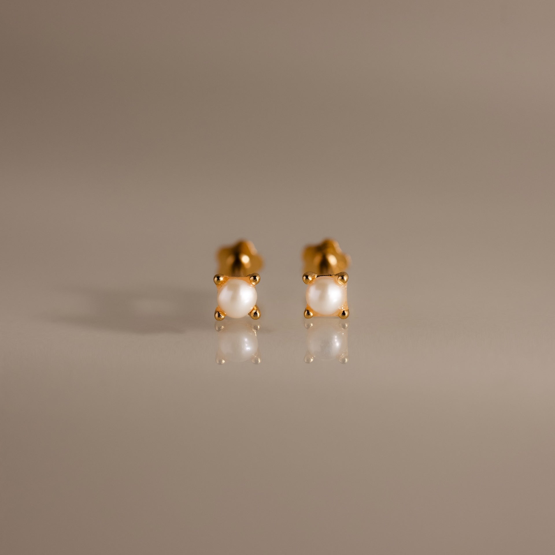A pair of Baby Venus Flatback Studs in 18K Gold, featuring delicate pearls and floral backing, displayed on a smooth, reflective surface.
