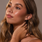 Woman with long brown hair wearing Baby Venus Flatback Studs in 18K Gold, looking upward and touching her neck against a neutral background.