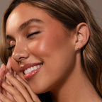 Woman with glowing skin smiles, eyes closed and hands near face, showcasing delicate Baby Venus Flatback Studs in 18K Gold with a subtle floral backing.