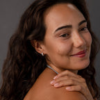 Smiling woman with wavy brown hair wears Ribbed Pave Huggies in Sterling Silver, touching her shoulder against a gray background.