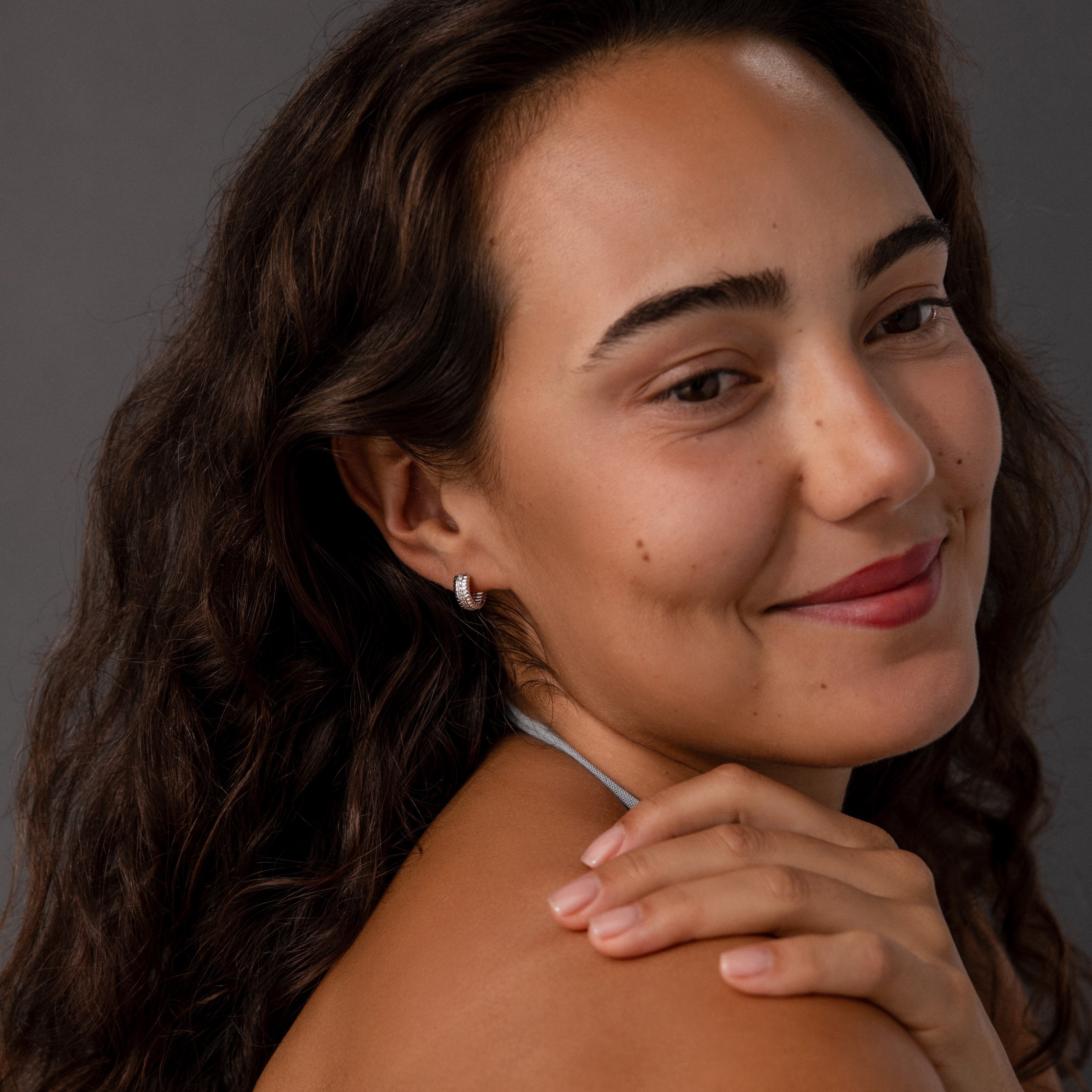 Smiling woman with wavy brown hair wears Ribbed Pave Huggies in Sterling Silver, touching her shoulder against a gray background.